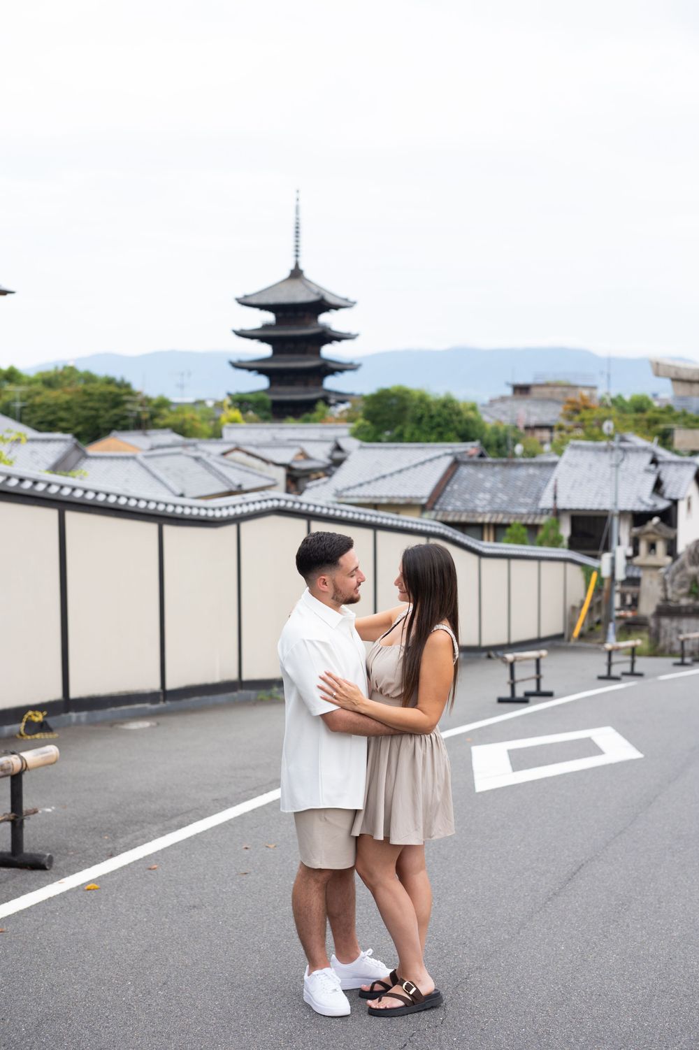 Couple embracing on a quiet Kyoto street with the Yasaka Pagoda, traditional rooftops and distant mountains in the background.