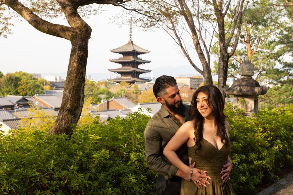 Couple embracing in a lush garden with a historic multi-tiered pagoda and stone lantern overlooking the city in soft golden light.