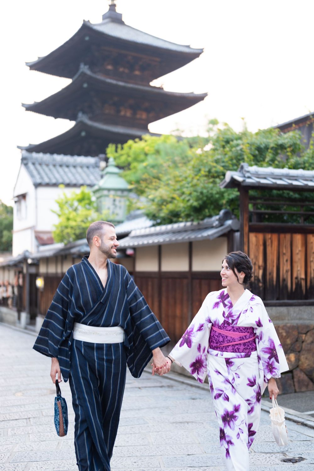 Smiling couple in traditional Japanese yukata holding hands while strolling past a historic wooden pagoda in Kyoto.