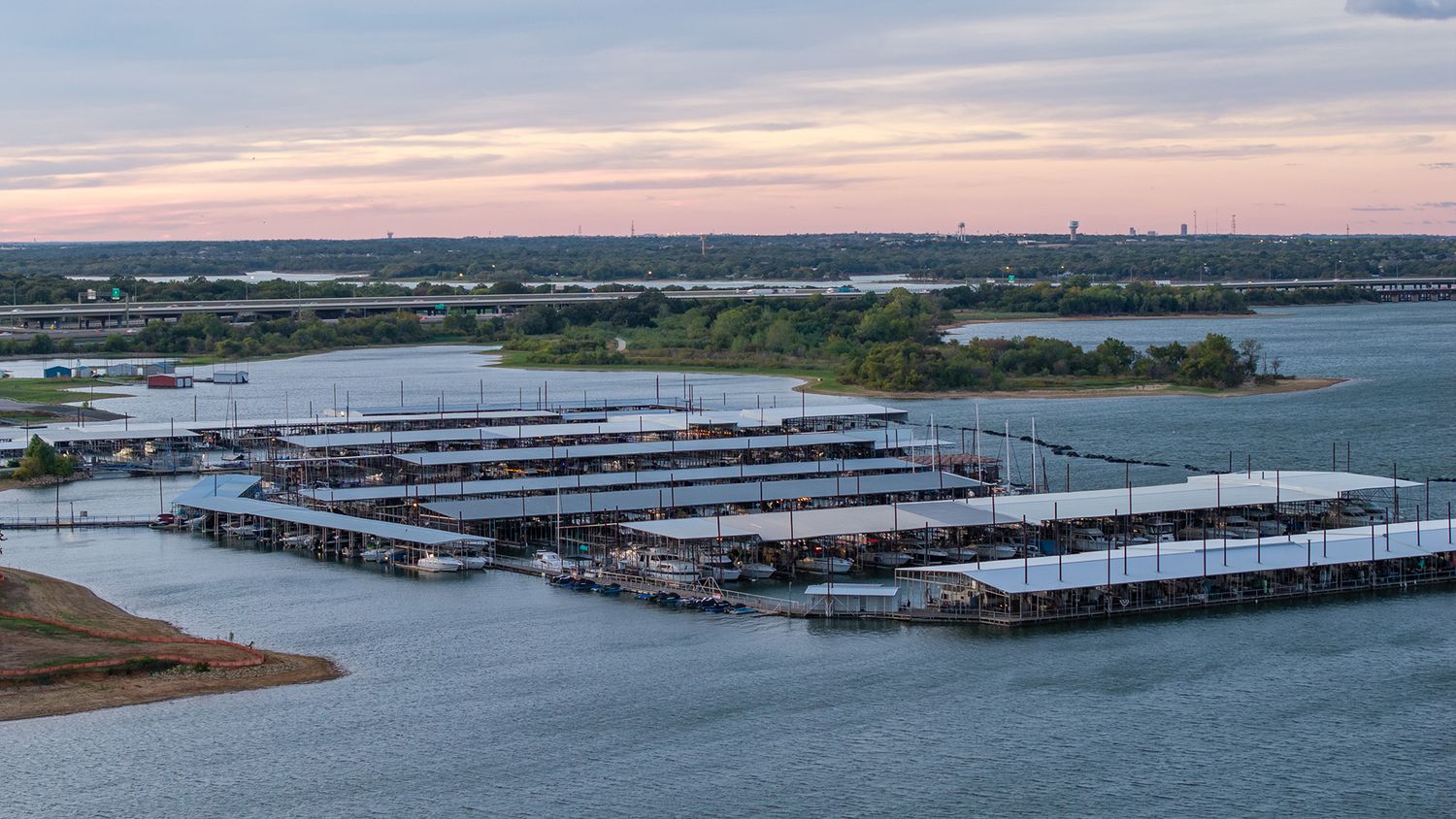 Aerial view of a lakeside marina with covered boat slips and docks along calm water at sunset, with trees lining the distant shore.