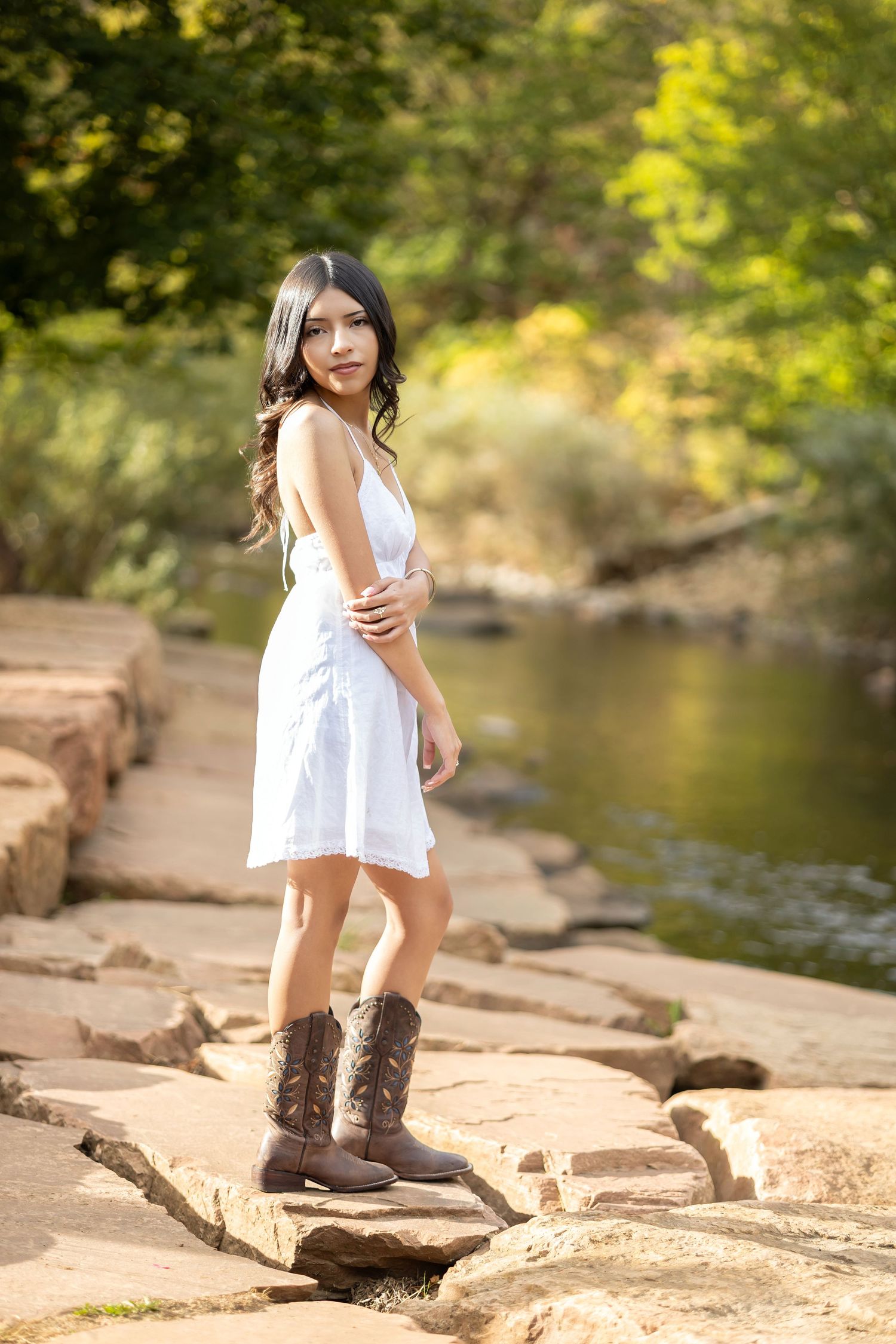 Young woman in white dress and cowboy boots standing on rocks near a serene creek surrounded by lush green trees.