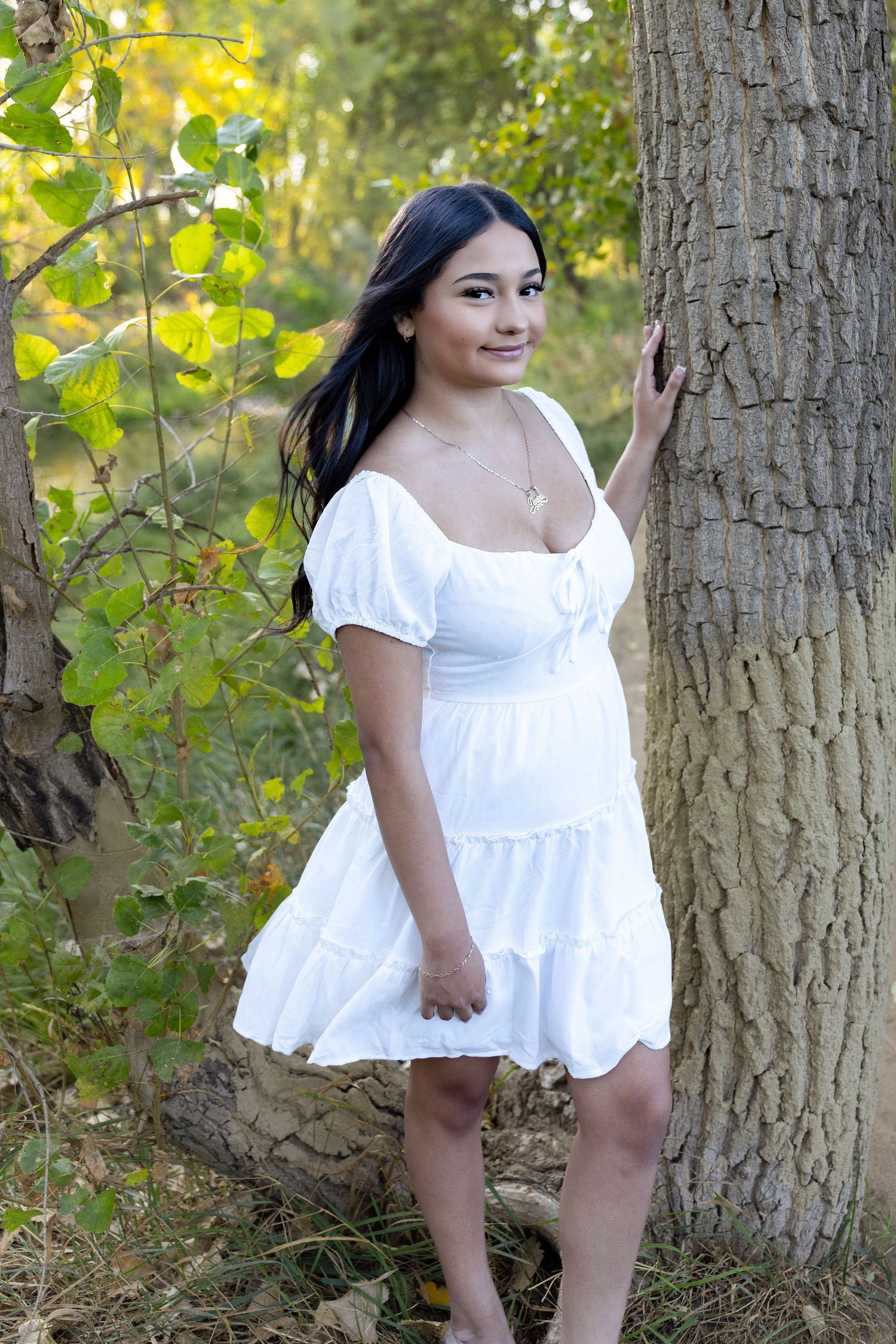 Young woman in white puff-sleeve mini dress poses by a tree in a lush green forest setting.