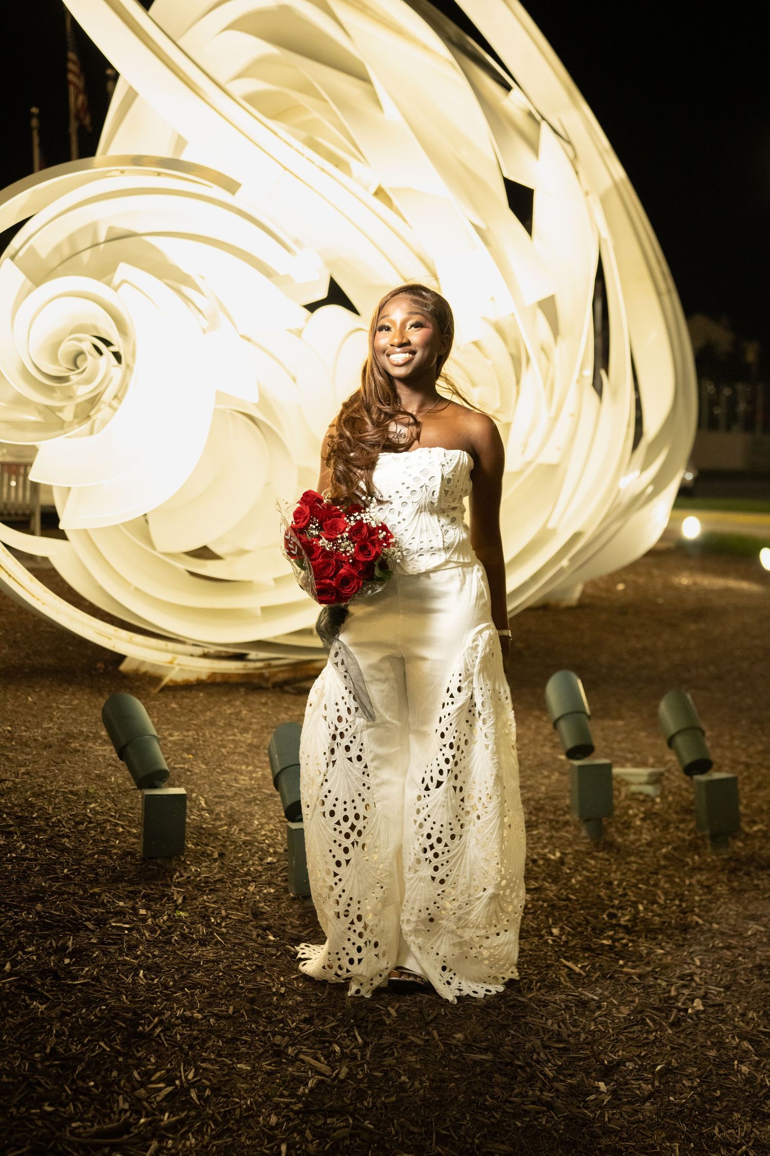 Smiling bride in white strapless gown holds red bouquet, posing outdoors at night with glowing light art behind her.