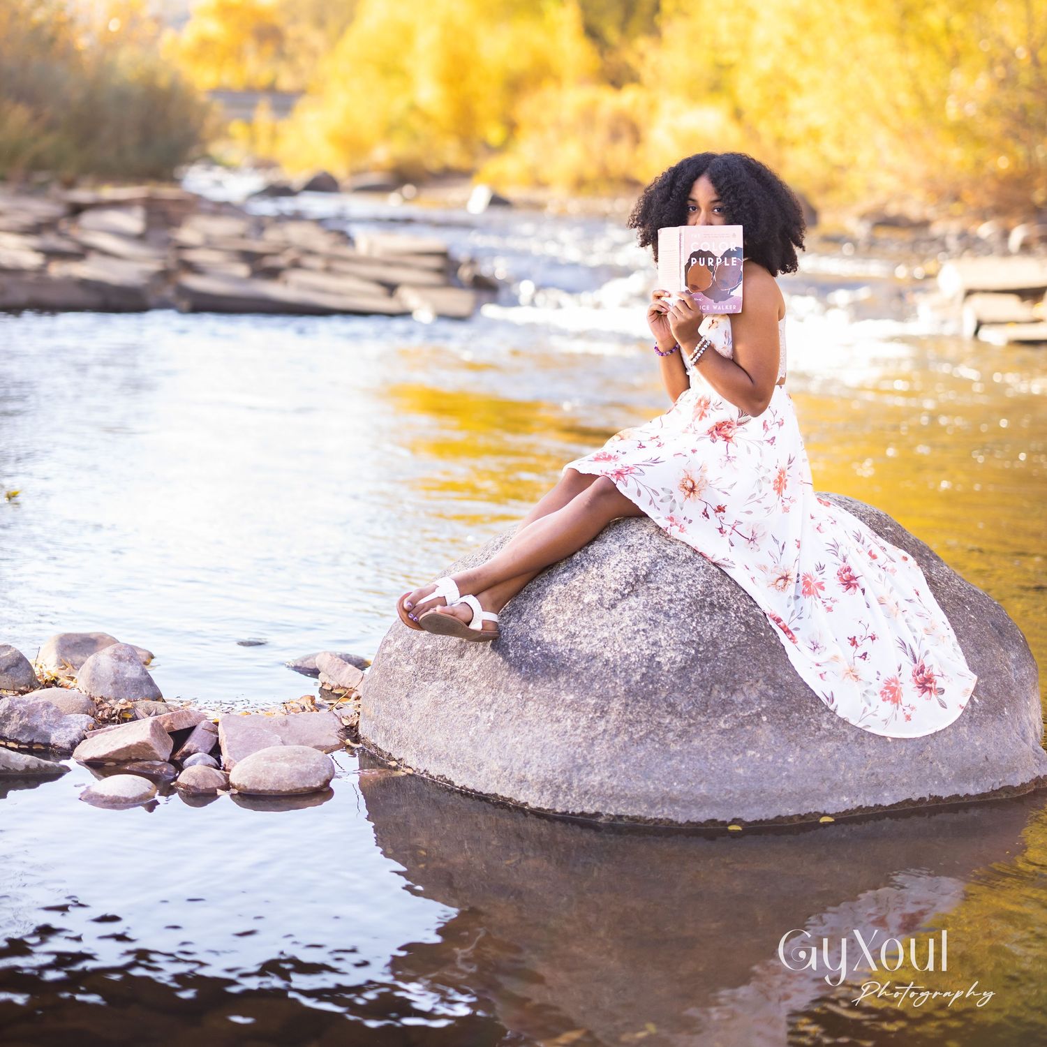 A young Black woman in a floral dress sits on a rock by a river, reading a book outdoors.