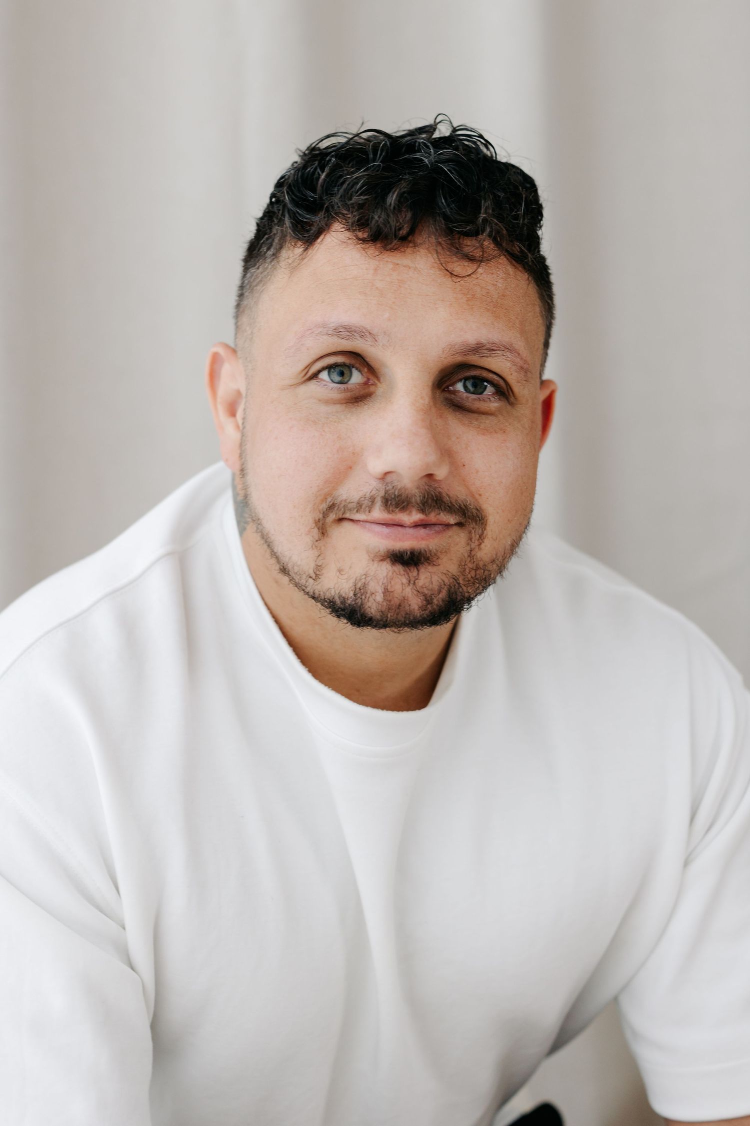 A man with curly dark hair and a beard wearing a white t-shirt, smiling softly against a light background.