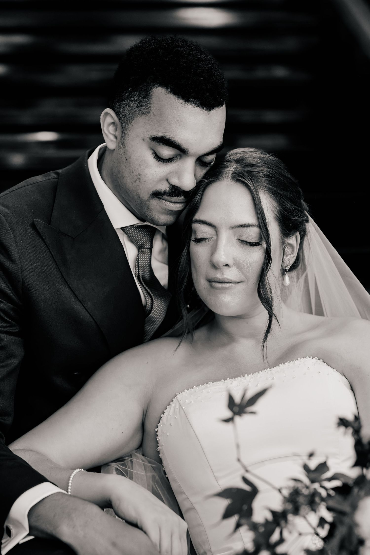 Black and white wedding portrait of a groom in a suit embracing a bride in a strapless gown and veil, both with eyes closed.