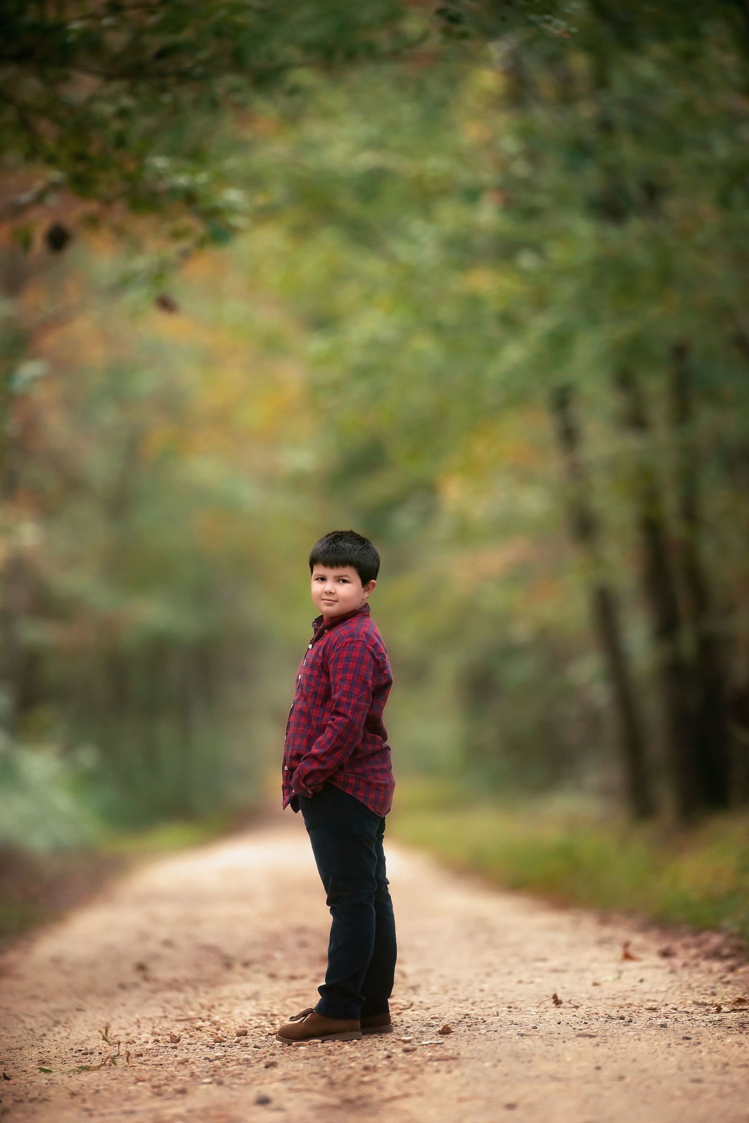 Young boy in a plaid shirt and dark jeans stands on a dirt road lined with autumn trees, glancing over his shoulder.