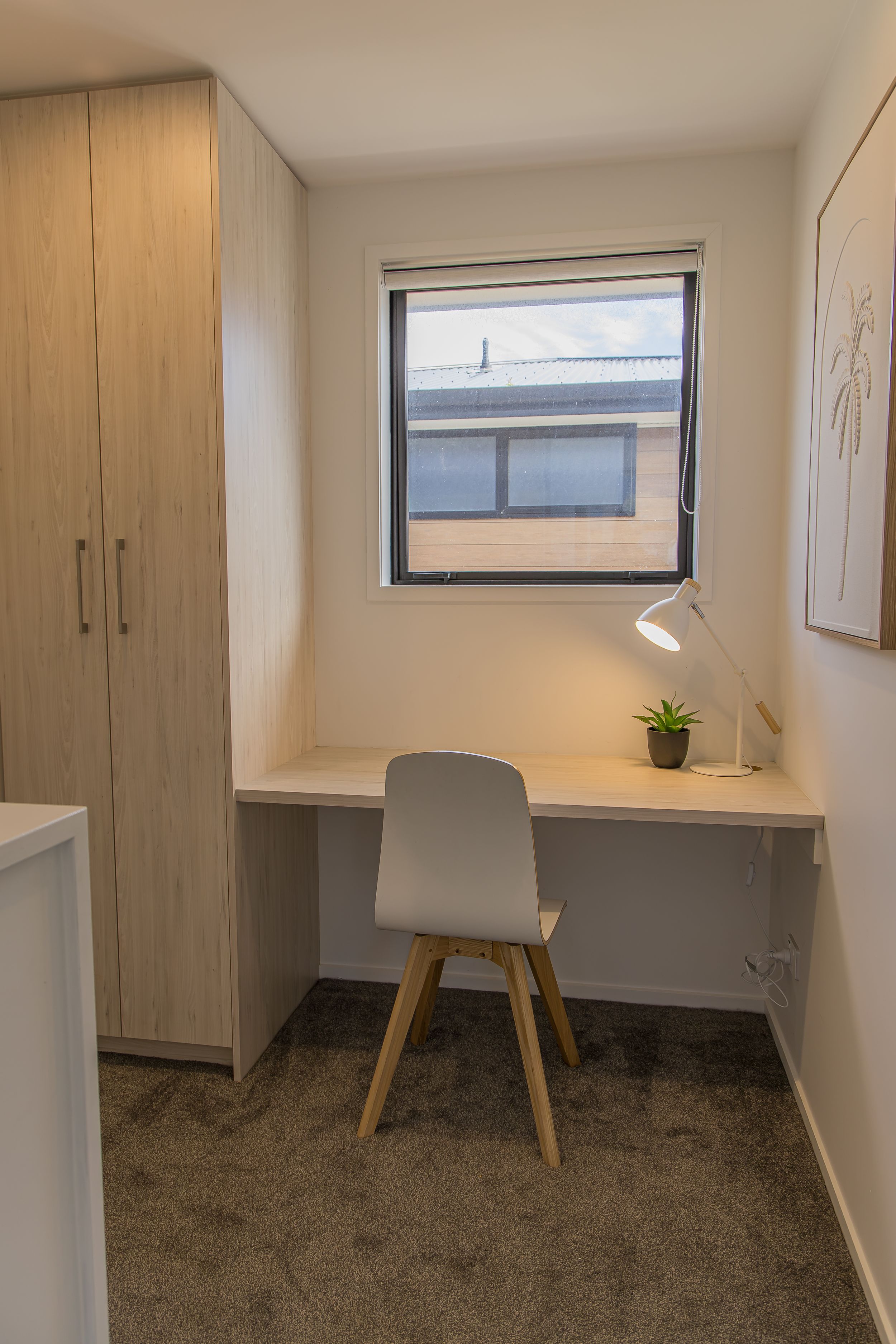 Compact home office nook with a built-in wooden desk, white modern chair, desk lamp and small plant beside a window and tall wardrobe.