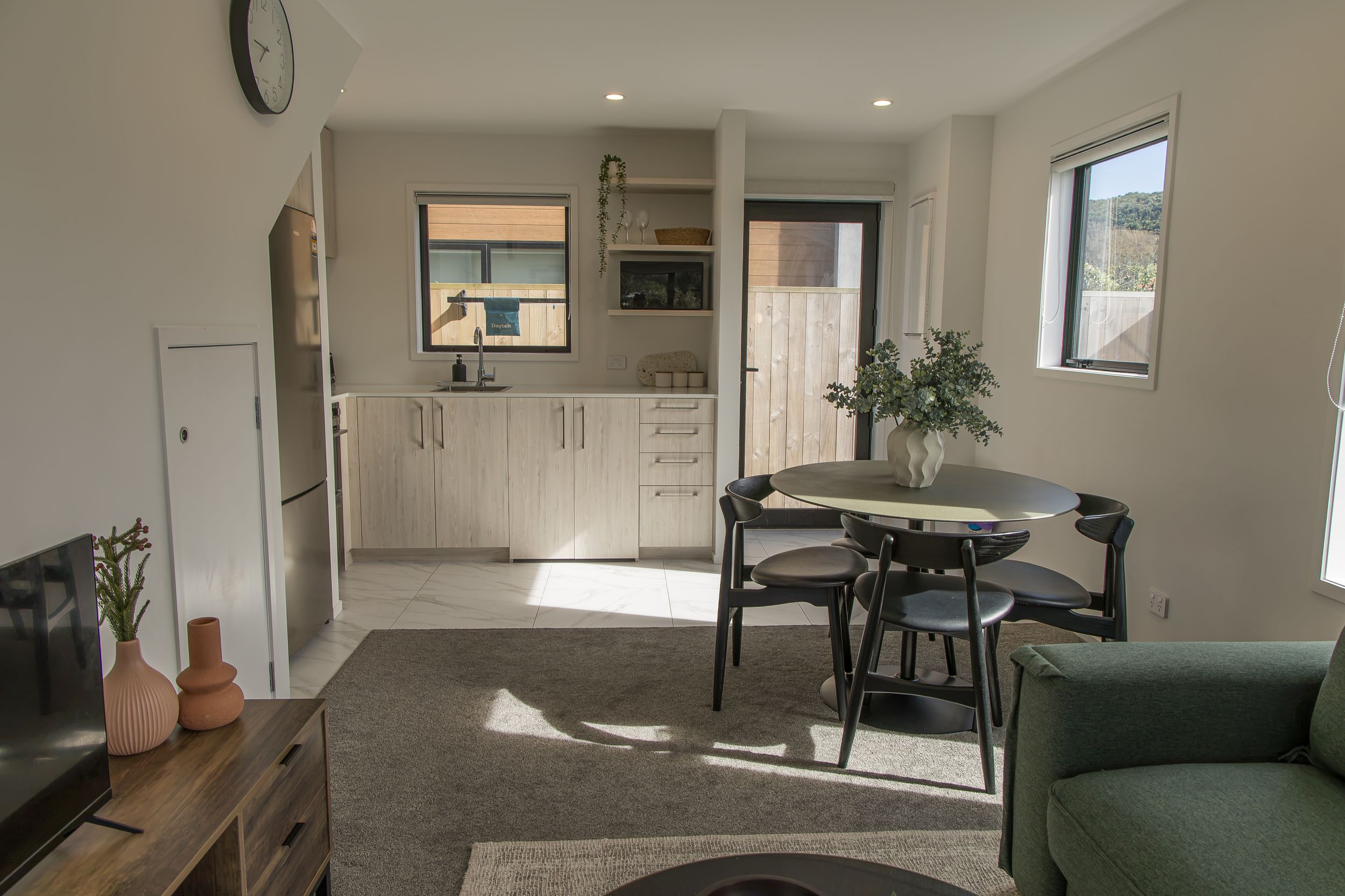 Bright open-plan living area with a round dining table and black chairs, a green sofa, wooden sideboard, and a compact kitchen beyond.