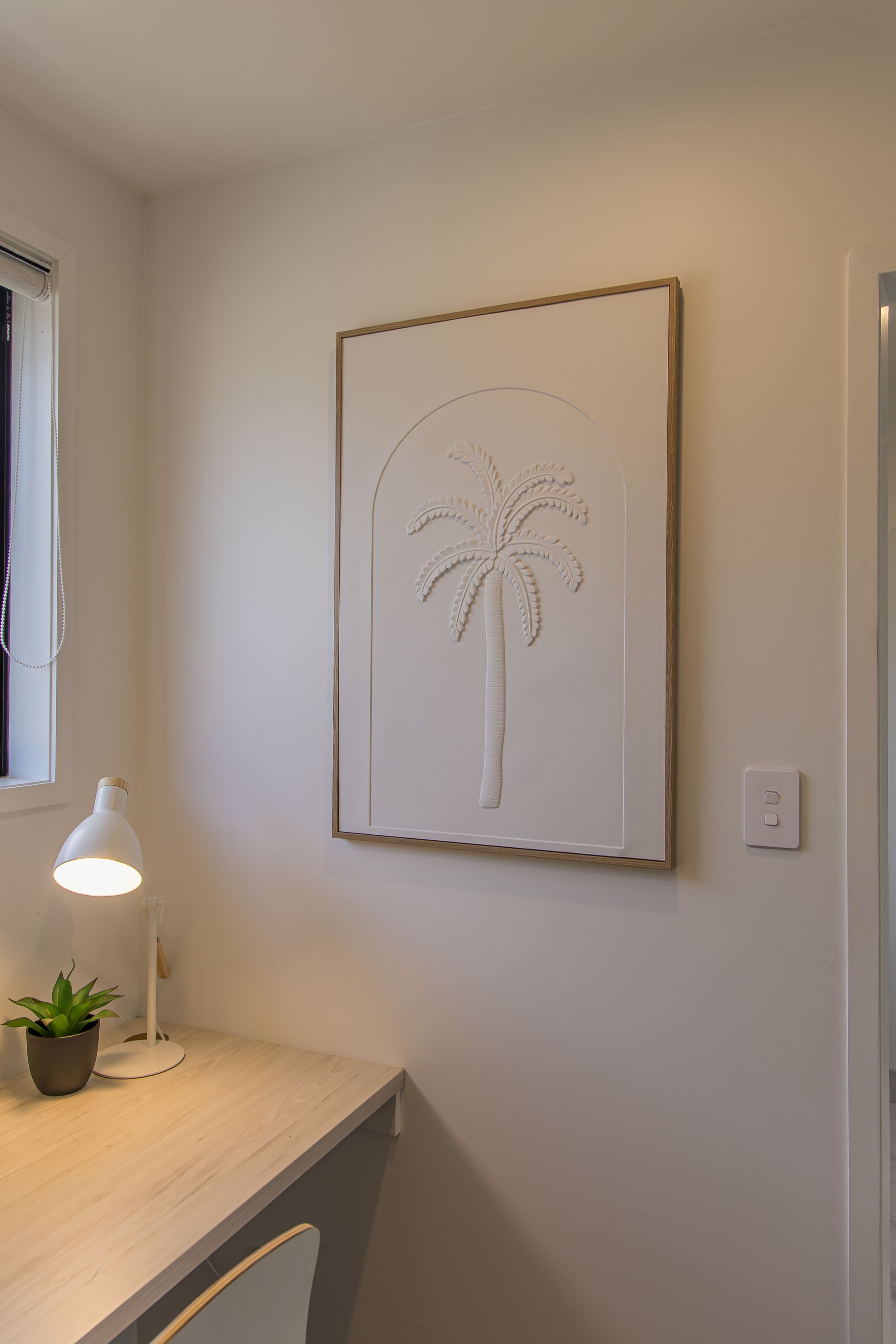 Minimalist workspace with a white palm tree relief artwork in a wooden frame above a desk with a white lamp and small succulent.
