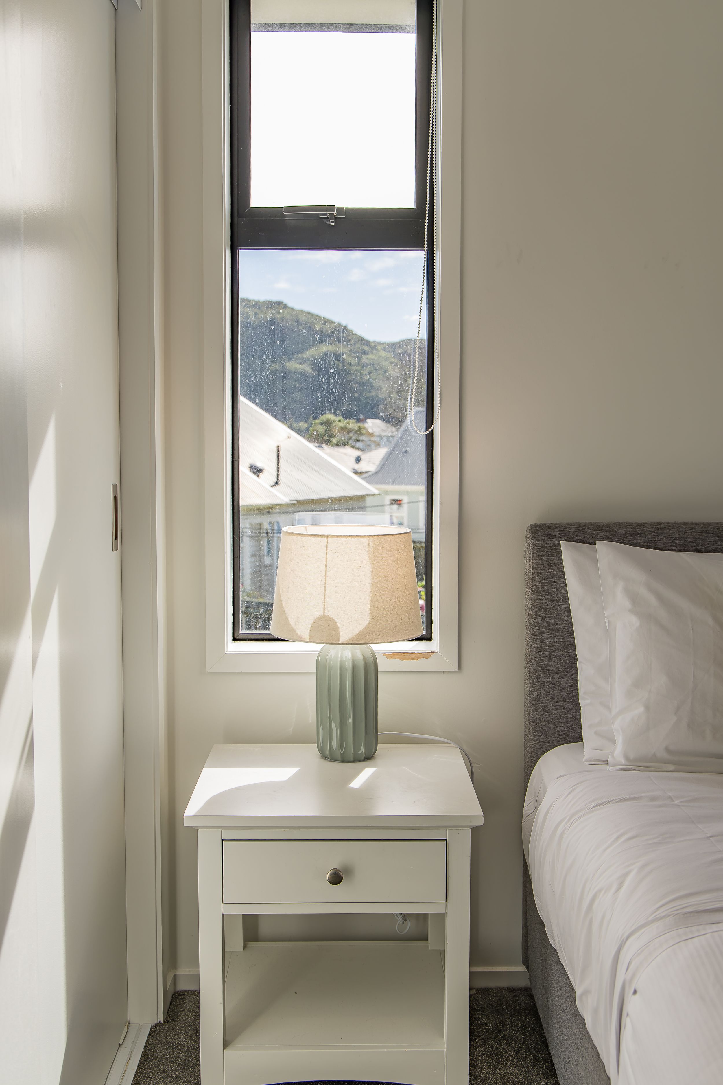 Bedroom corner with a white nightstand, sage green lamp and a tall window overlooking rooftops and a mountain view.