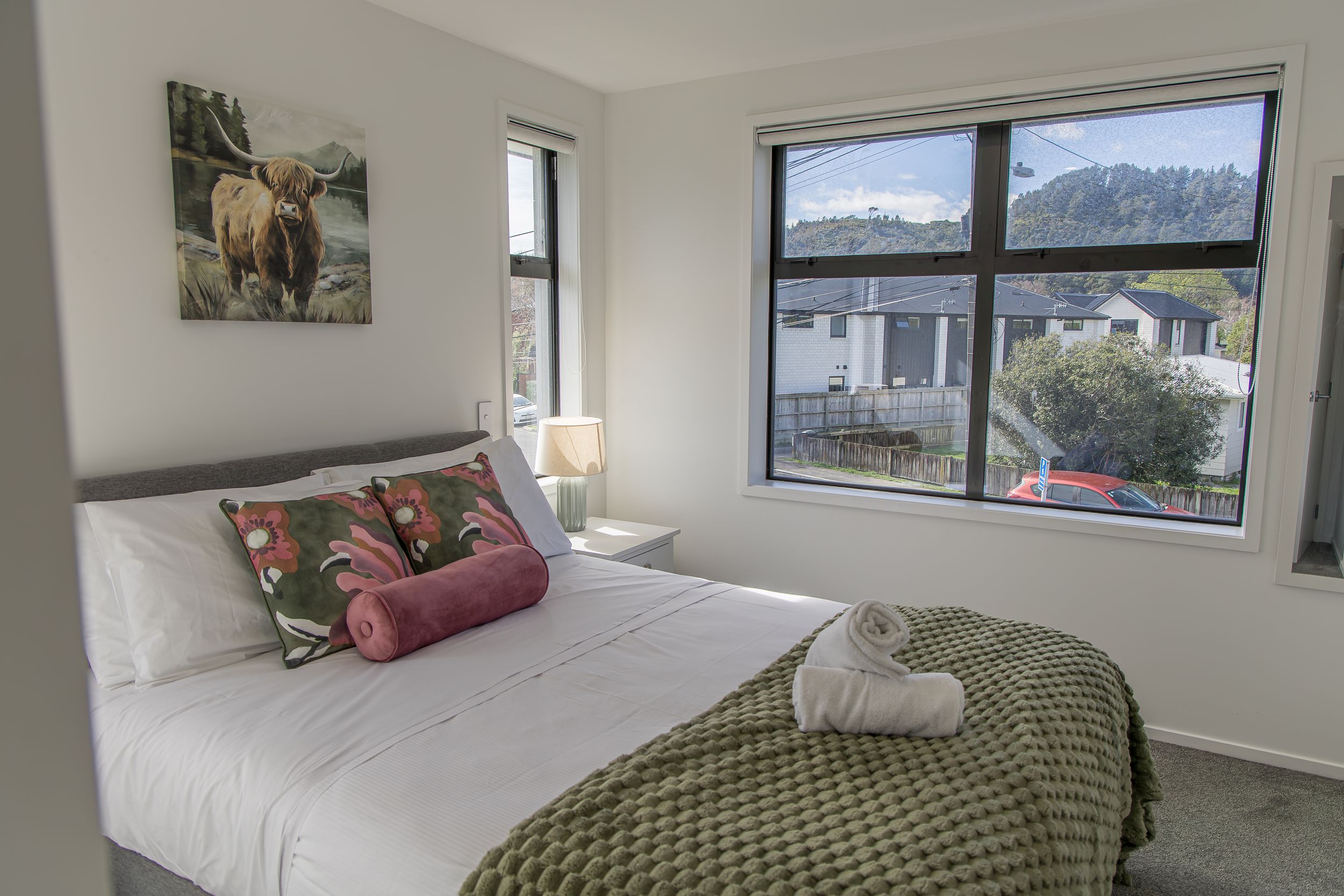 Bright modern bedroom with a double bed, green knitted throw, floral cushions, highland cow artwork and a large window with hillside views.