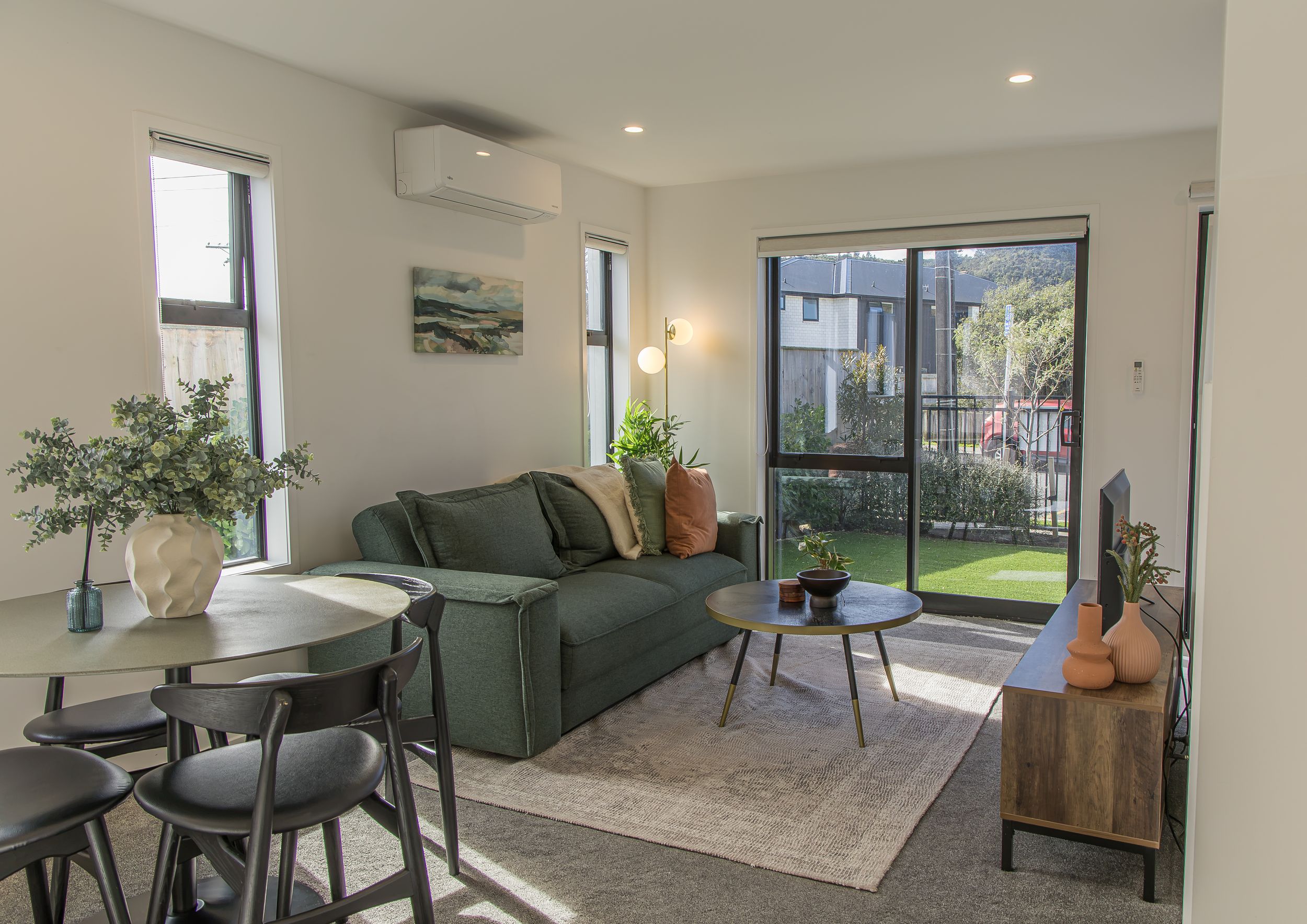 Bright open-plan living room with a green velvet sofa, round coffee table, dining set and glass doors opening to a sunny backyard.