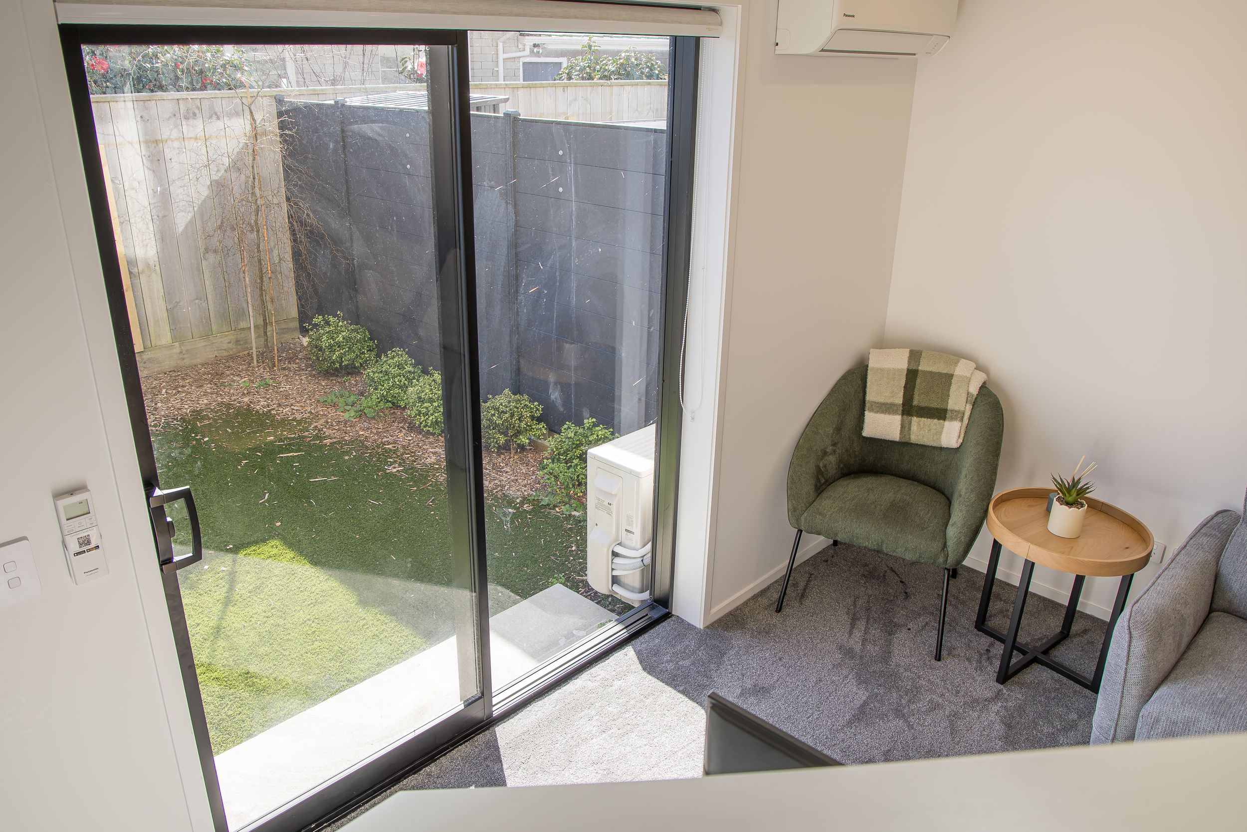 Sunlit living area with sliding glass door opening to a grassy fenced backyard, a green armchair and round wooden side table beside it.