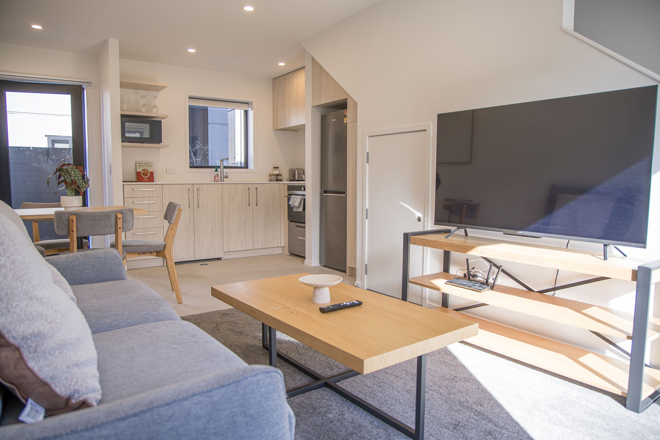 Open-plan apartment living area with a grey sofa, wooden coffee table, wall-mounted flat-screen TV and a compact kitchen with dining table.