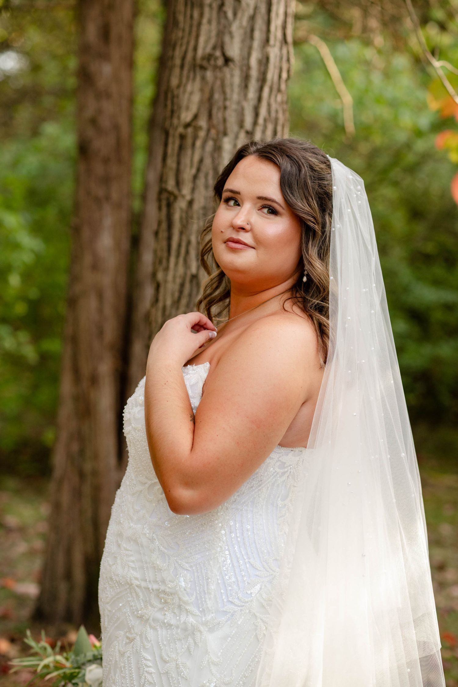 Bride in beaded strapless wedding gown with veil posing beside a large tree in a forest.