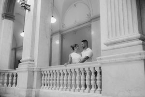 Black and white photo of a wedding couple standing between marble columns on an ornate balcony.