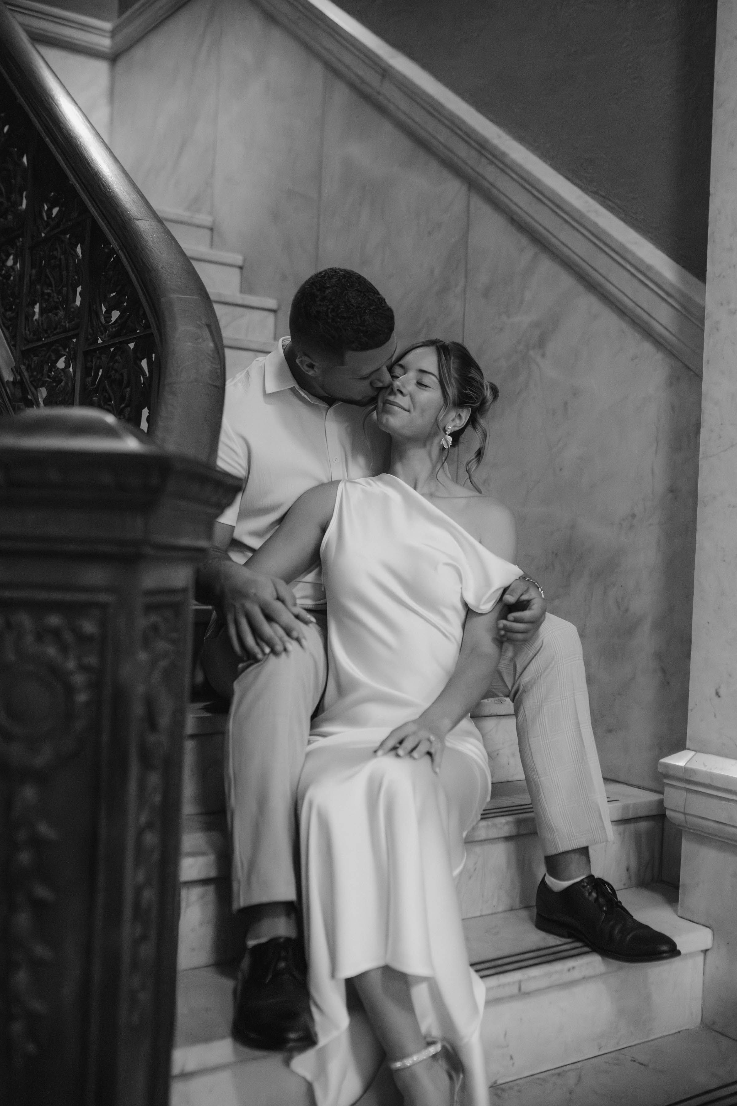 Black and white romantic photo of a couple sitting on marble stairs, sharing a tender kiss.