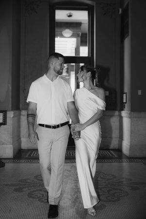 Black and white photo of a newlywed couple walking hand in hand through an elegant building hallway.