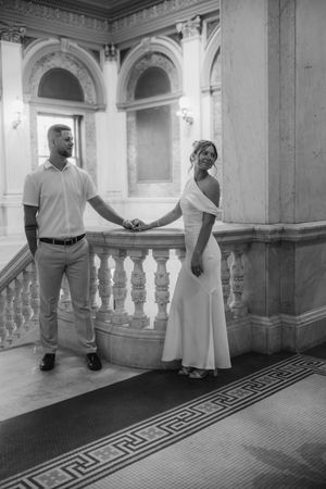 Black and white photo of a wedding couple holding hands on ornate marble stairs in a grand building.