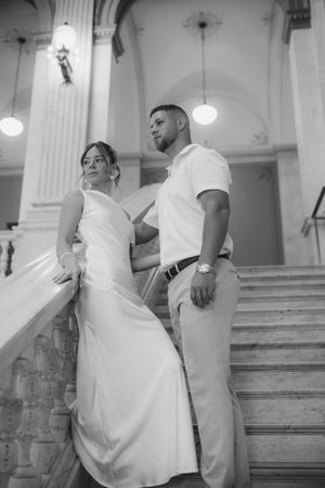 Black and white photo of bride and groom posing elegantly on marble staircase in grand historic building.