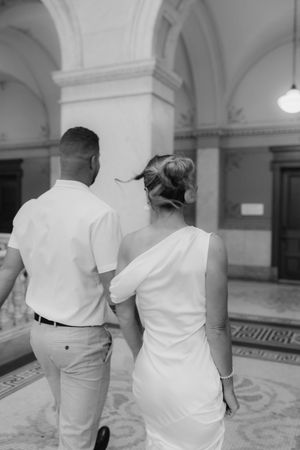 Black and white rear view of bride and groom walking hand in hand through grand arched hallway.