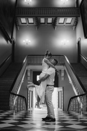 Black and white photo of groom lifting bride in romantic embrace at base of grand double staircase.
