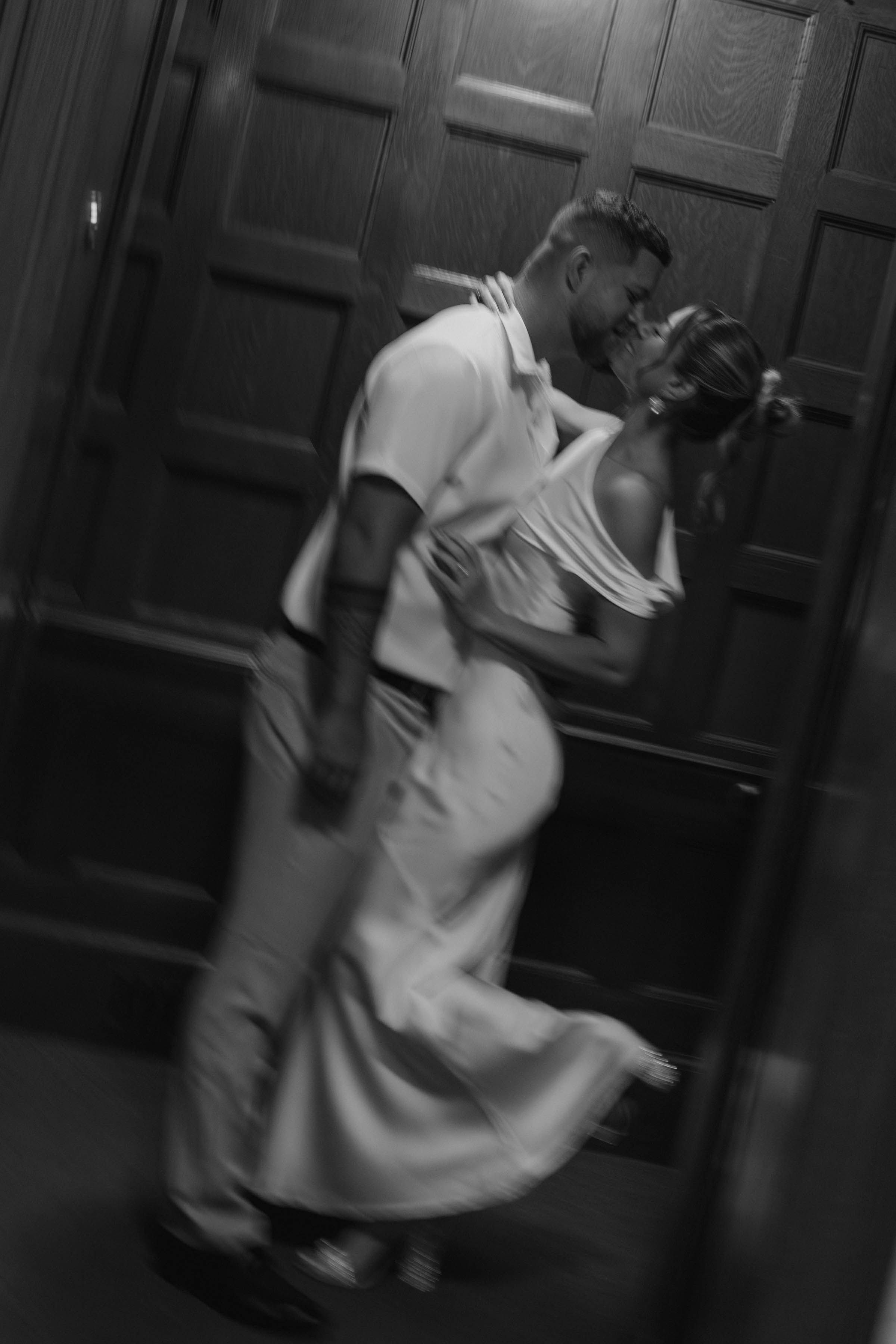 Blurred black and white artistic photo of couple kissing and dancing near wooden doors.