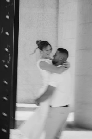 Blurred black and white artistic photo of groom lifting smiling bride between stone columns.