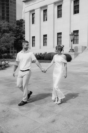 Black and white photo of bride and groom holding hands while walking outside historic building.