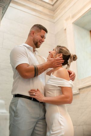 Groom gently cups brides face as they share an intimate moment in a marble building.