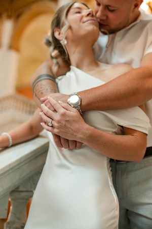 Close-up of groom embracing bride from behind showing wedding rings and watch details.