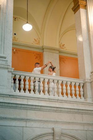 Groom twirls bride on ornate marble balcony of a stunning classical courthouse building.