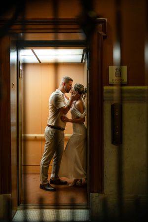 Newlyweds sharing an intimate embrace inside a vintage wooden elevator with warm golden lighting.