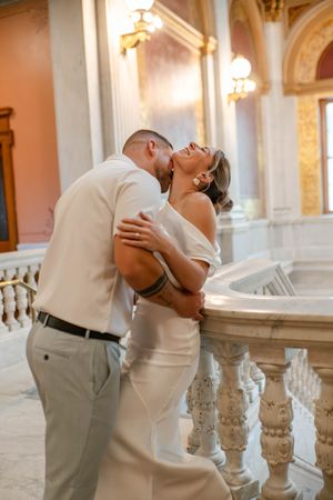 Groom kissing bride's neck as she laughs joyfully near ornate marble balustrade in elegant venue.