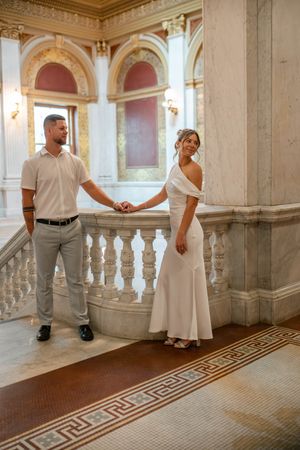 Bride and groom holding hands near ornate marble staircase railing in a grand classical building.