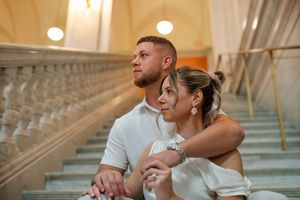 Couple sitting together on marble stairs, bride in white off-shoulder gown gazing thoughtfully ahead.