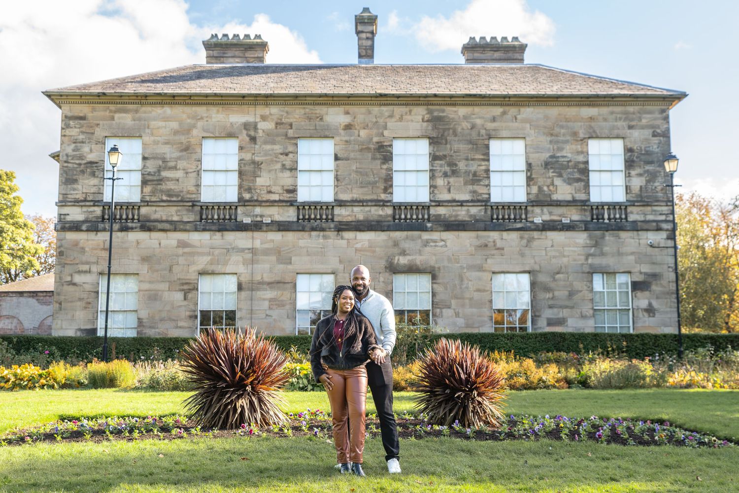 Couple embracing in front of a grand historic stone mansion with manicured gardens on a sunny day at Clifton Park. Engagement photography