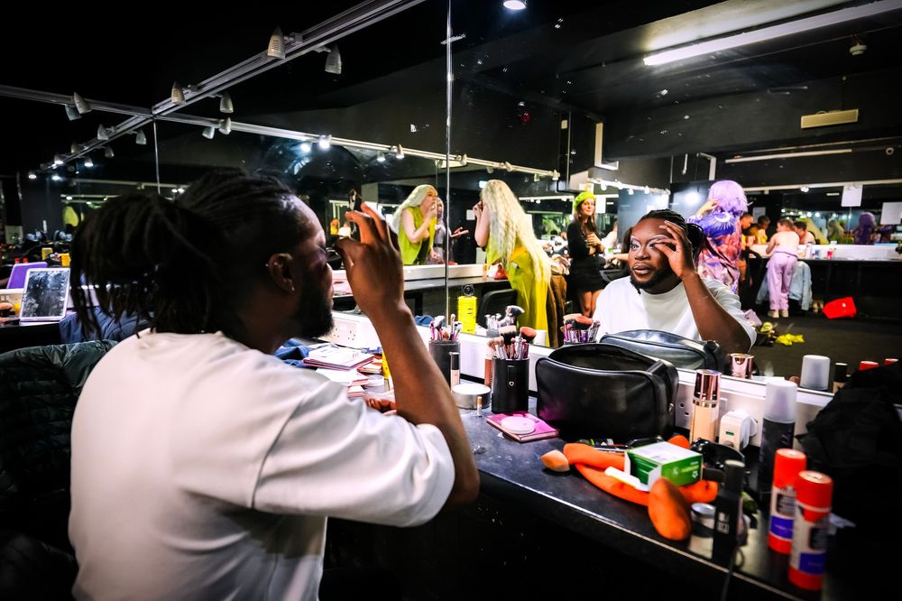 Two performers apply makeup at a backstage mirror surrounded by beauty products under dramatic lighting.