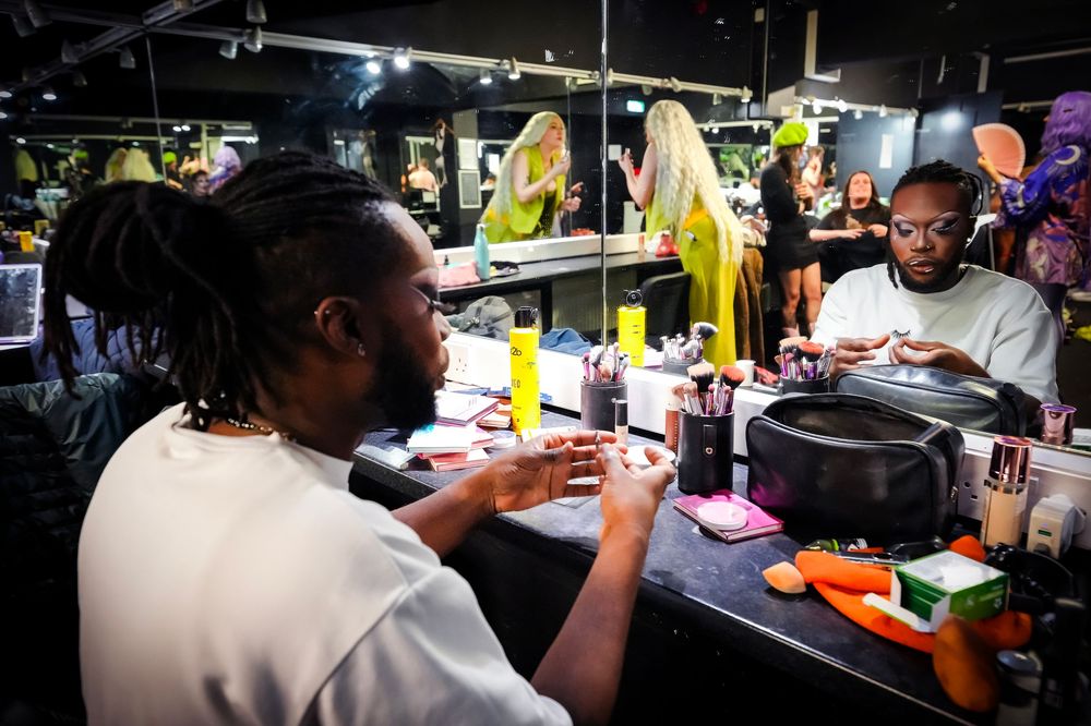 Two men sit at a backstage makeup mirror surrounded by colorful products and bright lights.