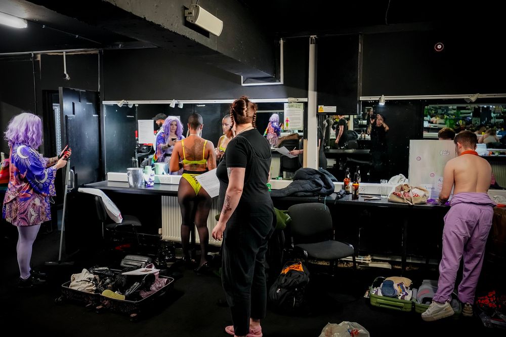 Backstage dressing room with performers in colorful costumes preparing in front of lit mirrors.