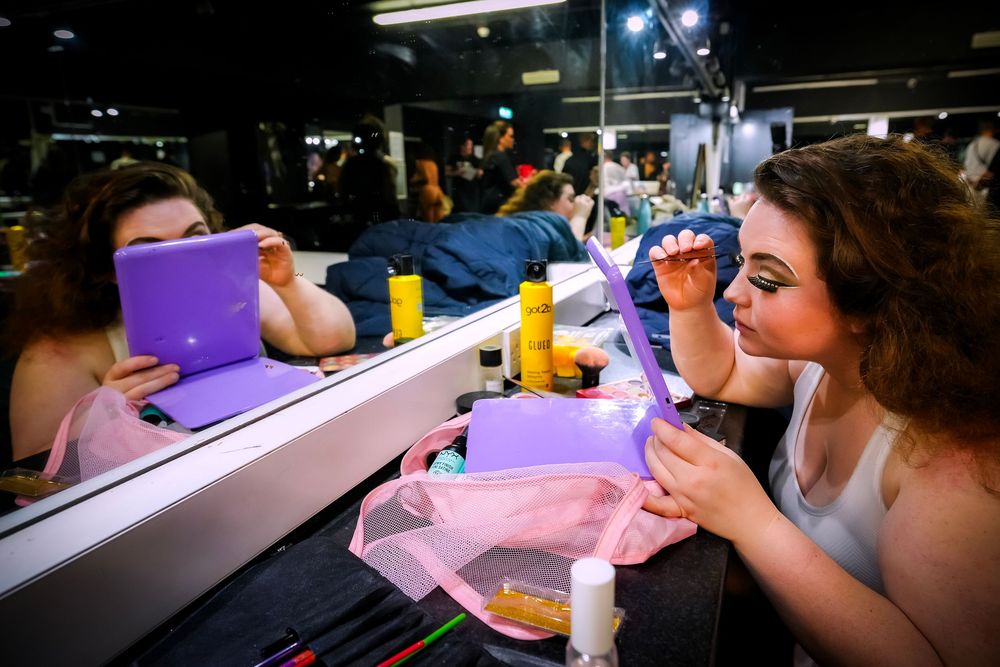 Young woman applying makeup at a vanity mirror backstage, surrounded by beauty products and a purple tablet.