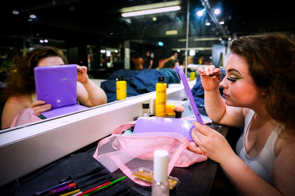 Young woman applying makeup at a vanity mirror with colorful beauty products on the table.