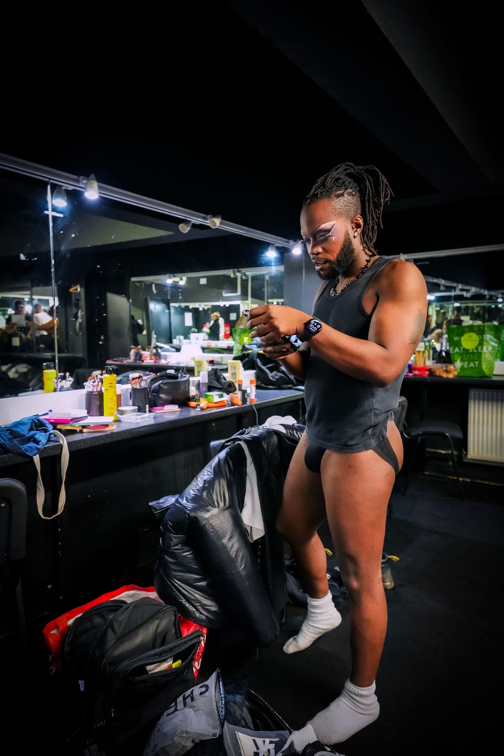 Muscular athlete in black bodysuit checks phone in dimly lit backstage dressing room with mirrors and bags.