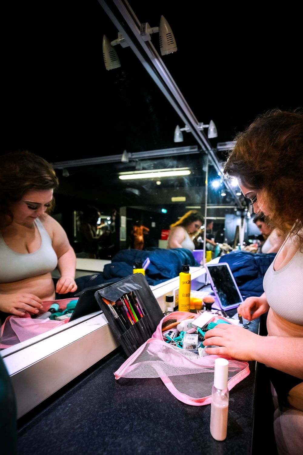 Woman applies makeup at a dressing room mirror with lights, surrounded by cosmetics and beauty tools backstage.