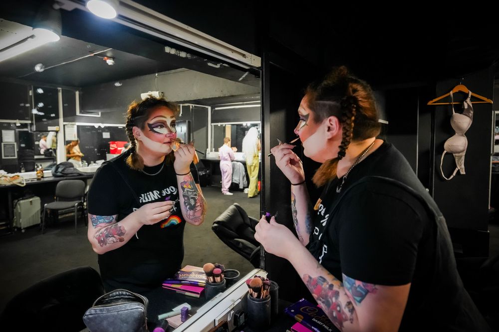 Two performers apply dramatic stage makeup in a dressing room mirror before a show.