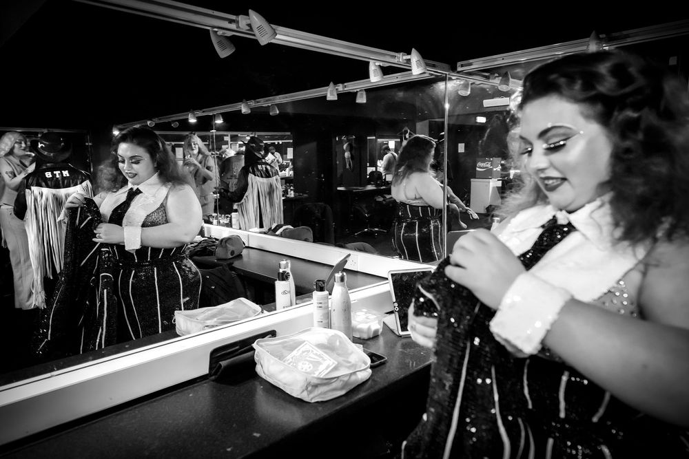 Black and white photo of a woman in costume applying makeup at a dressing room mirror backstage.