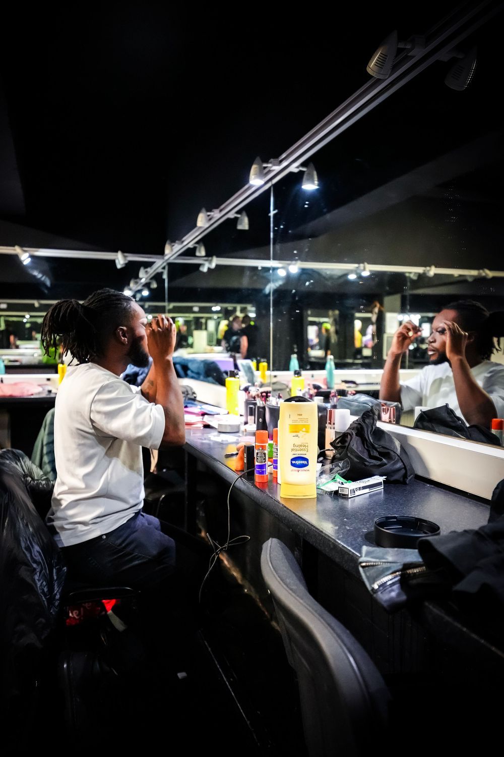 Man in white shirt applies makeup at a dimly lit dressing room mirror with beauty products on the counter.