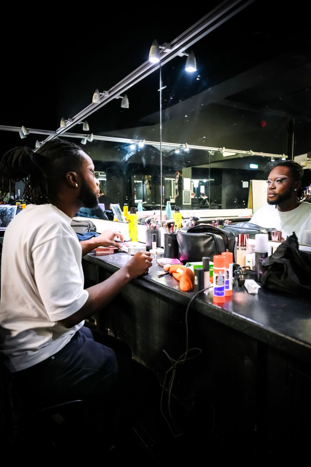 Man sitting at a barbershop vanity mirror with grooming products, lit by overhead lamp in dark salon.