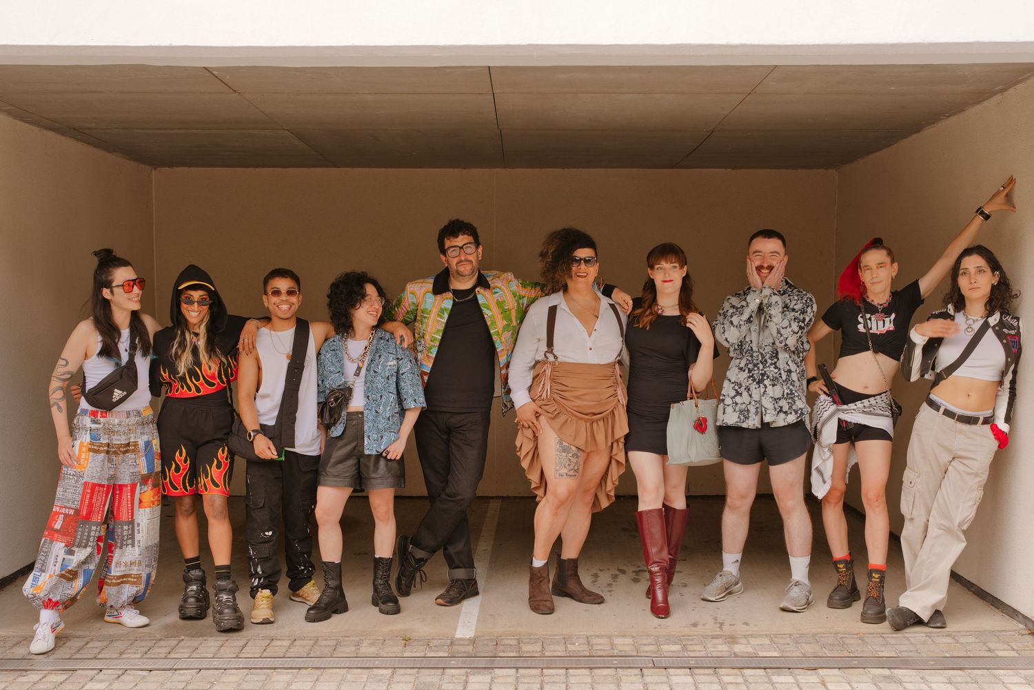 Group of young adults in colorful festival costumes posing together under a covered outdoor walkway on a sunny day.